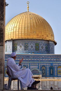 a man sitting on a chair in front of the dome of the rock with his hand up