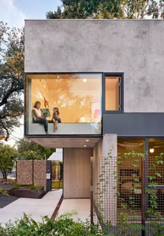 two people sitting on the window sill of a modern house with trees in the background