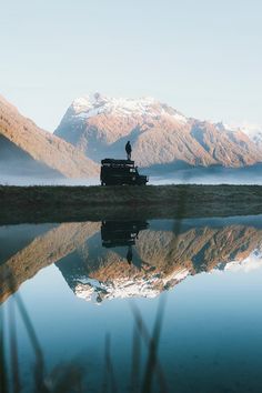 a man standing on top of a truck next to a lake with mountains in the background