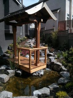 a wooden gazebo sitting next to a small pond in front of a house with rocks on the ground