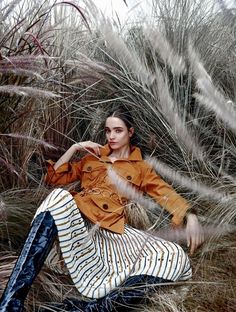 a woman sitting in tall grass with her hand on her hip, wearing an orange jacket and striped skirt