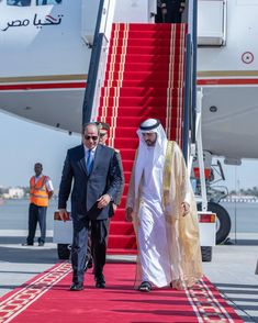 two men in suits walking down a red carpeted walkway next to a white plane