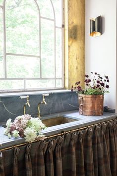 a kitchen sink under a window next to a counter with flowers on it and a potted plant