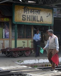 two men are standing on the train tracks in front of a railway station with an old sign that says shimla