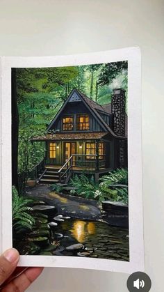 a hand holding an open book with a painting of a cabin in the woods on it