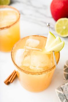 two glasses filled with drinks and garnished with apples, cinnamon sticks and lime