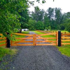 a wooden gate in the middle of a gravel road leading to a grassy field with trees