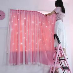 a woman standing on a pink ladder next to a window covered in sheer curtains and lights