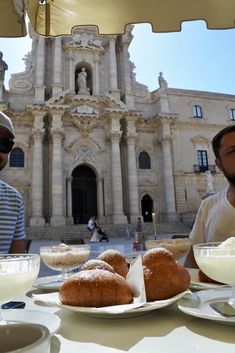 two men sitting at a table with plates of food and drinks in front of an old building