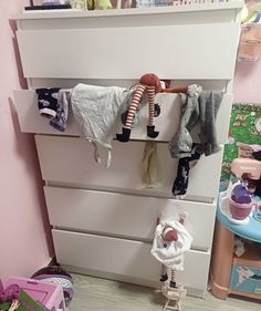 a white dresser with clothes hanging on it's top and two children's toys in the bottom drawer