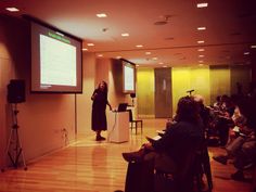 a woman standing in front of a projection screen giving a presentation to people sitting on chairs