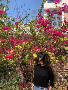 a woman standing in front of a bush with pink flowers on it and looking at the ground