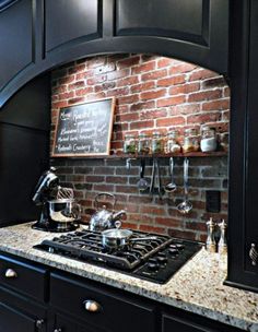 a kitchen with black cabinets and brick wall behind the stove top is filled with cooking utensils