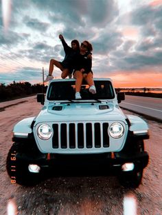 two people sitting on the hood of a jeep with their arms in the air and one person standing on top