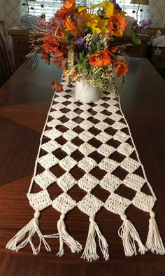 a crocheted table runner with tassels and flowers in the vase on top