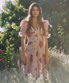 a woman standing in the middle of a field with flowers on her dress and hair blowing in the wind