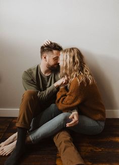 a man and woman sitting next to each other on top of a wooden floor in front of a white wall