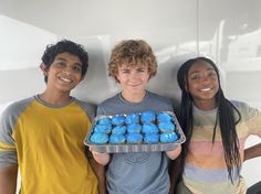 three young people holding a tray of blue cupcakes