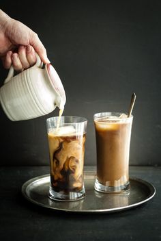 a person pours coffee into two glasses on a silver tray with another cup in the background