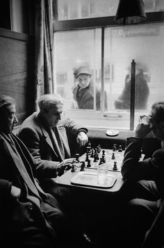 black and white photograph of men playing chess in front of a window with people looking out the window