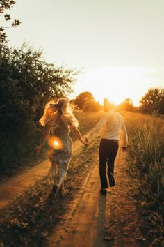 a man and woman walking down a dirt road holding hands as the sun goes down