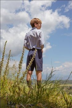 a young man standing on top of a lush green field next to tall grass and clouds