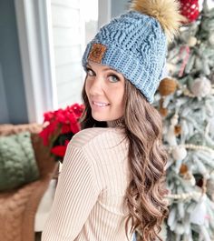 a woman standing in front of a christmas tree wearing a blue hat with pom - poms