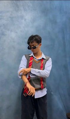 a young man wearing sunglasses and a vest standing in front of a blue wall with his arms crossed