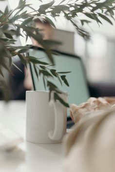 a white coffee cup sitting on top of a table next to a laptop computer and a plant