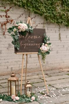 a welcome sign with flowers and greenery on the ground next to a brick wall