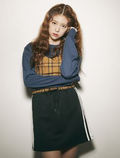 a girl with long red hair wearing a blue sweater and black skirt standing in front of a white wall