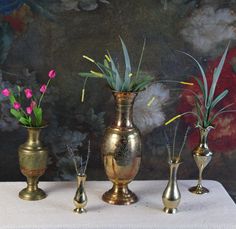 three brass vases with flowers in them on a white table cloth next to a painting