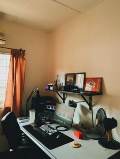 a desk with a laptop computer on top of it next to a fan and window