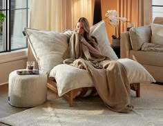 a woman wrapped in a blanket is sitting on a couch with pillows and a foot stool