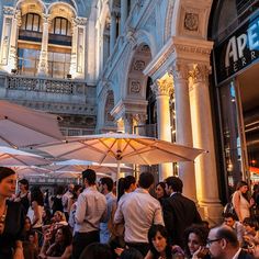 a group of people sitting at tables under umbrellas in front of a large building