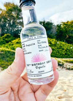 a person holding up a bottle of birthday gin on the beach with trees in the background