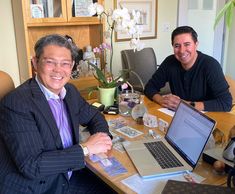 two men sitting at a table with laptops and flowers in the vase behind them