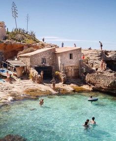 people are swimming in the clear blue water near some rocks and buildings on top of a cliff