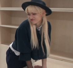 a blonde woman with long hair wearing a black hat and blue shirt is leaning on a shelf