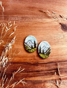 two buttons with trees and mountains on them sitting on a wooden table next to some dry grass