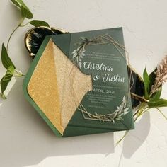 a green and gold wedding stationery on top of a table next to some greenery