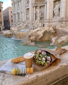 a cup of coffee and some food on a ledge next to a fountain with statues