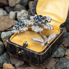 This vintage floral brooch features two (2) flowers with leaves and a stem pavé and partially bezel set with sixty-seven (67) round brilliant cut diamonds. The flowers are centered with a total of twenty-three (23) prong set, round mixed cut sapphires. The stem is channel set with seven (7) baguette cut diamonds. The brooch measures 49.1mm long by 37.8mm wide and 14.2mm thick, finished with a hinged and locking pin back. One of the round diamonds has a small chip. Diamond Flower Brooch For Formal Occasions, Formal Diamond Flower Brooch, Vintage Brooches With Brilliant Cut Diamonds, Flowers With Leaves, 2 Flowers, Floral Brooch, Baguette Cut Diamond, Channel Set, Baguette Cut