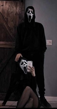 a person in a black outfit with a white mask on their face and holding a cell phone