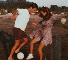 a man and woman kicking a soccer ball on the ground with trees in the background