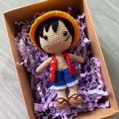 a crocheted doll in a box with purple shredded paper on the floor next to it