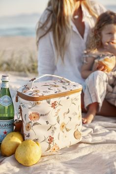 a mother and daughter sitting on the beach with lemons, water bottle and cooler bag