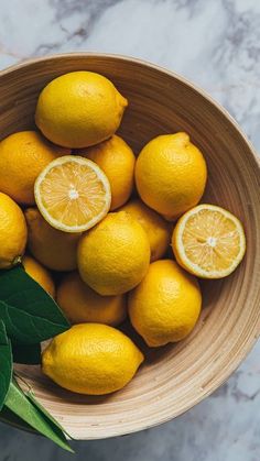 a wooden bowl filled with lemons on top of a marble counter next to a green leaf