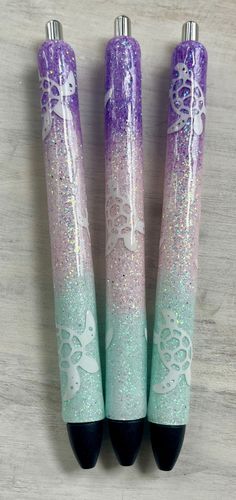 three purple and green pens sitting next to each other on a wooden table with glitter