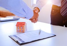 two people shaking hands over a document on a table with a house model and pen